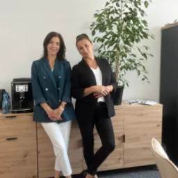 Dwie kobiety w biurowych strojach stojące obok drewnianej komody z ekspresem do kawy i rośliną doniczkową, prezentacja zespołu.