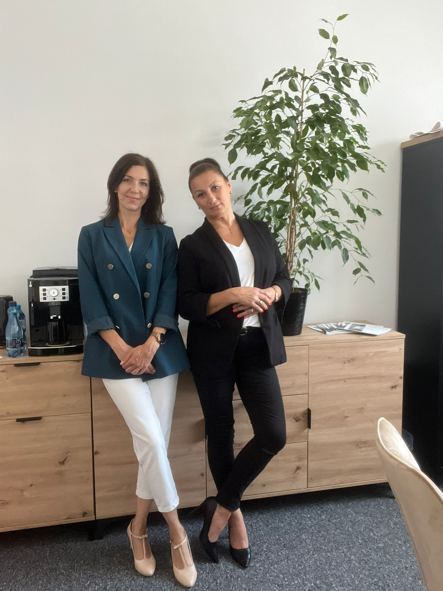 Dwie kobiety w biurowych strojach stojące obok drewnianej komody z ekspresem do kawy i rośliną doniczkową, prezentacja zespołu.