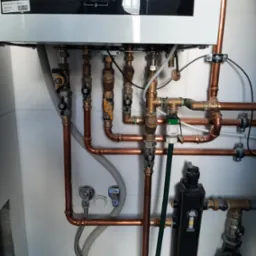 Nowoczesny piec gazowy kondensacyjny z widocznymi miedzianymi rurami, zaworami i filtrem wody, zamontowany na białej ścianie z płytkami.