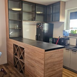 MebleParadise - Nowoczesna kuchnia z drewnianą wyspą z wbudowanym regałem na wino, przeszklonymi szafkami wiszącymi z czarnymi ramami, czarnym blatem i sprzętem AGD w tle.