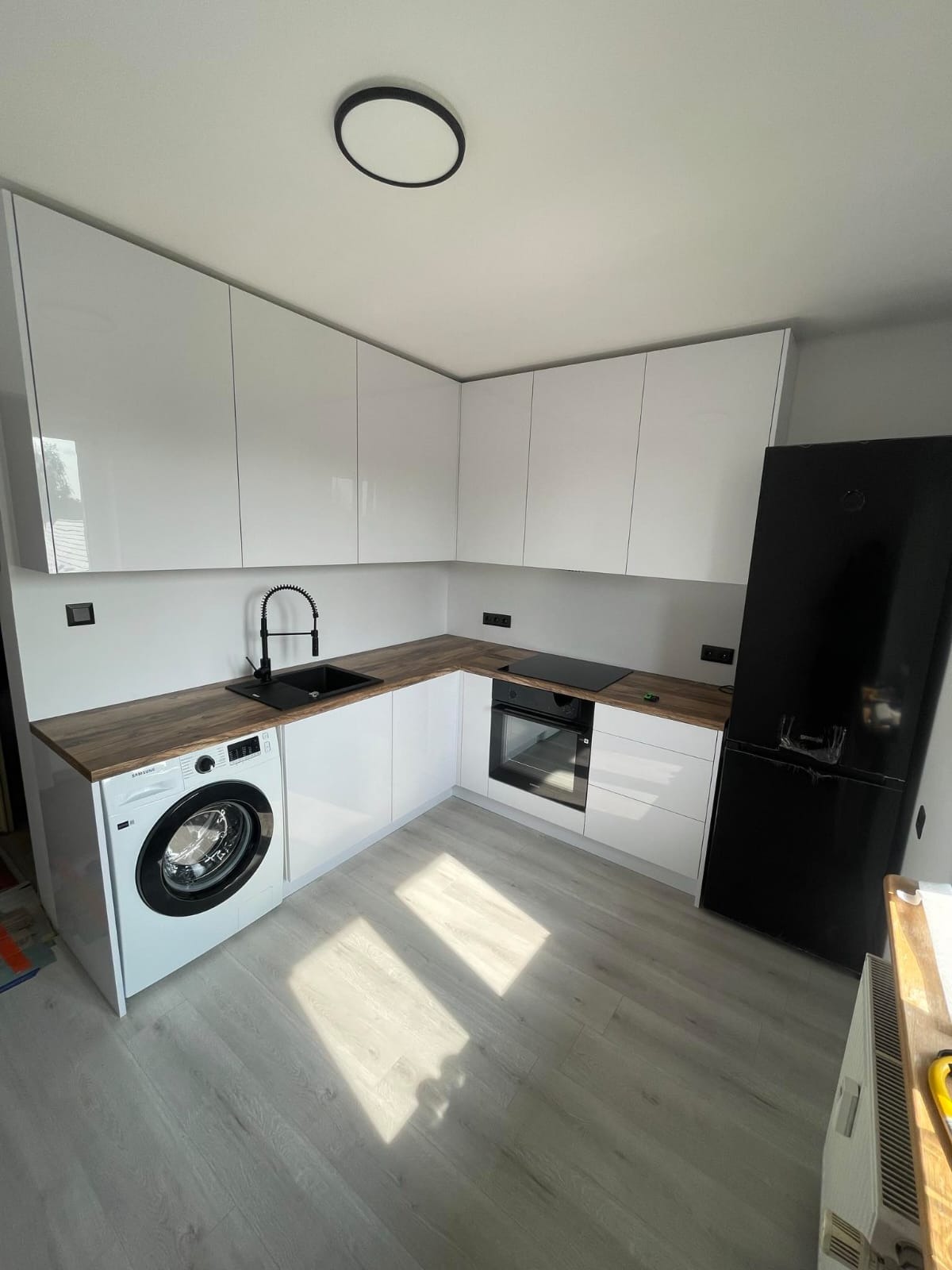 Biała, lakierowana zabudowa kuchenna w kształcie litery L z drewnianym blatem, czarnym zlewem i baterią, pralką w zabudowie oraz czarną lodówką.