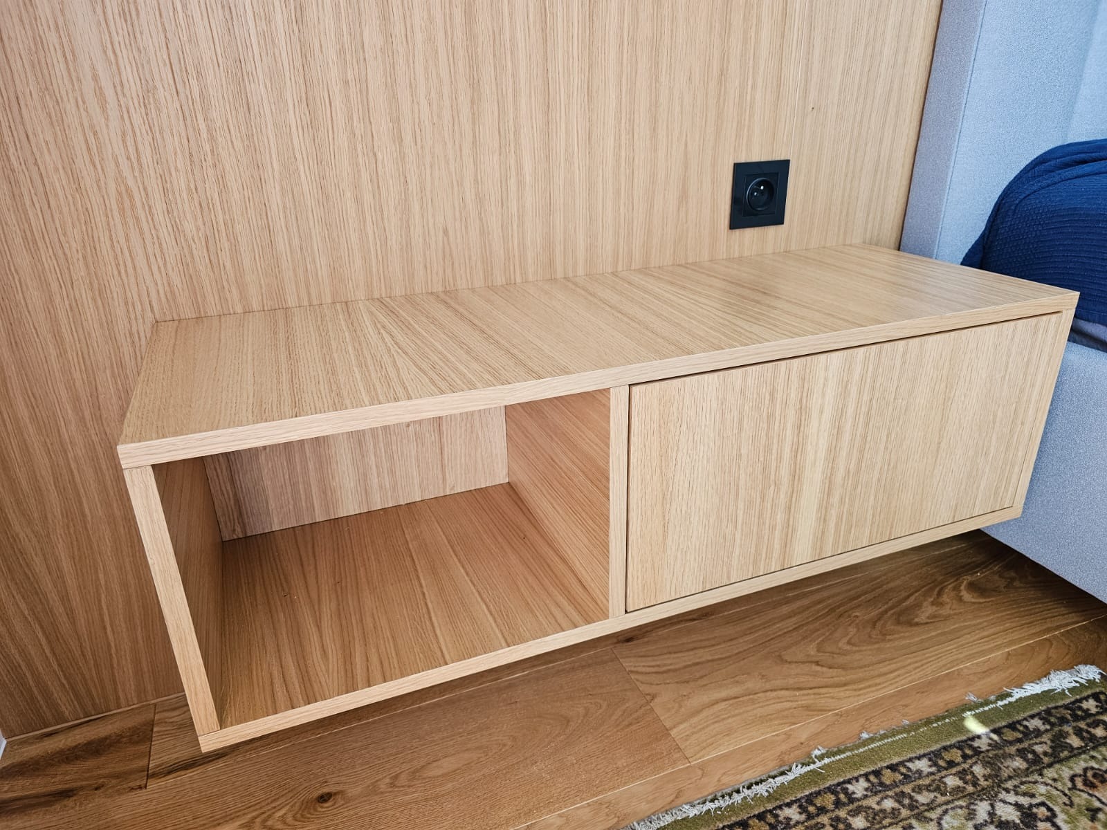 Drewniana szafka nocna z otwartą półką i zamykaną szafką, zamontowana na ścianie obok łóżka z widocznym gniazdkiem elektrycznym.