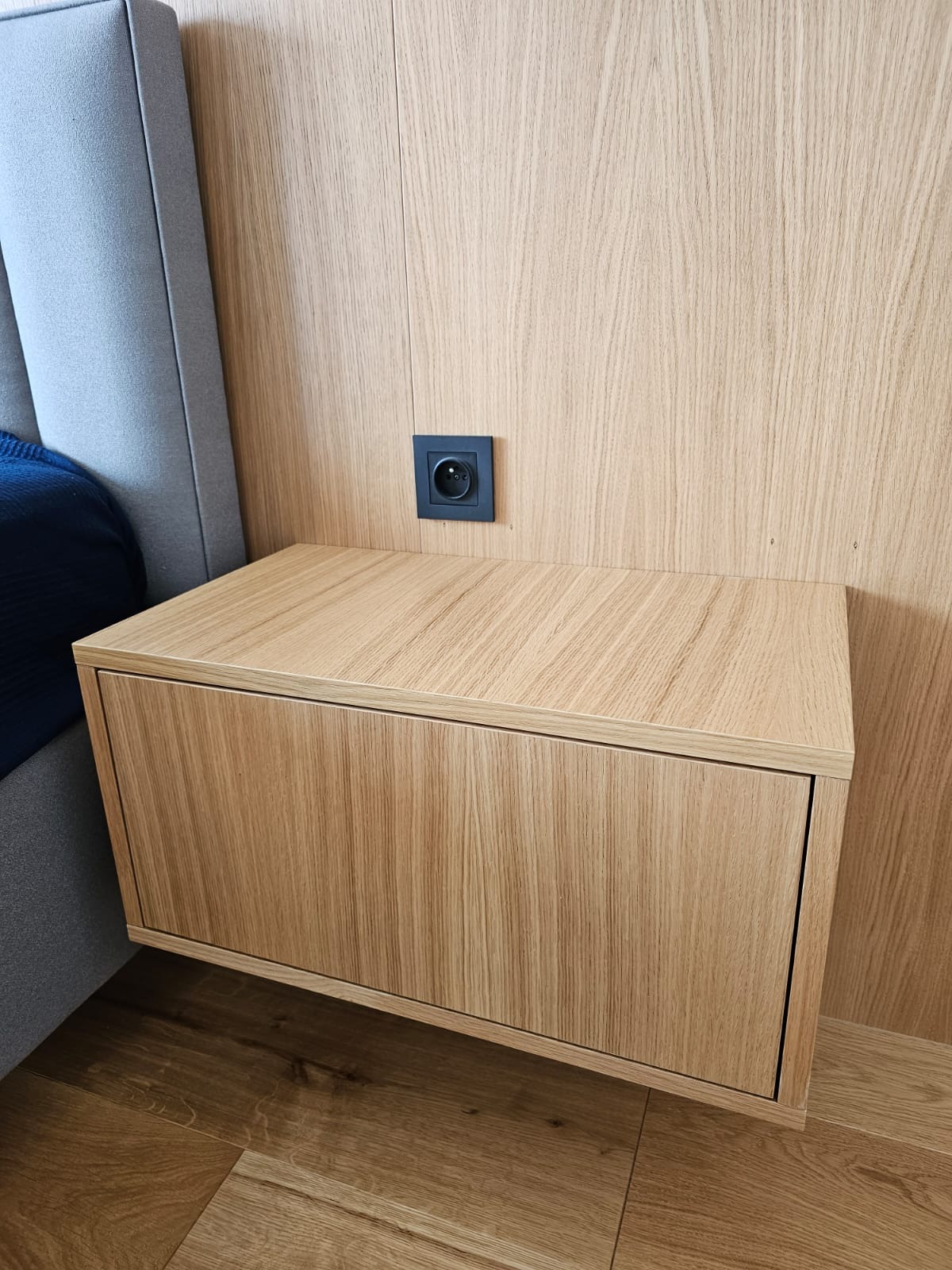 Drewniana szafka nocna z szufladą, wisząca na ścianie obok łóżka z szarą ramą, z czarnym gniazdkiem elektrycznym powyżej.