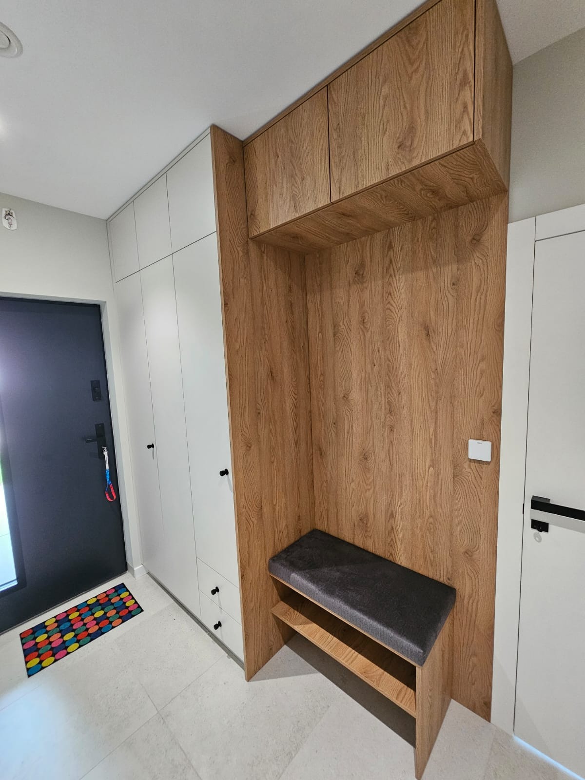 Wnęka z siedziskiem i szafkami z drewnopodobną okleiną, połączona z białą szafą w przedpokoju, widoczna wycieraczka w kolorowe kropki.
