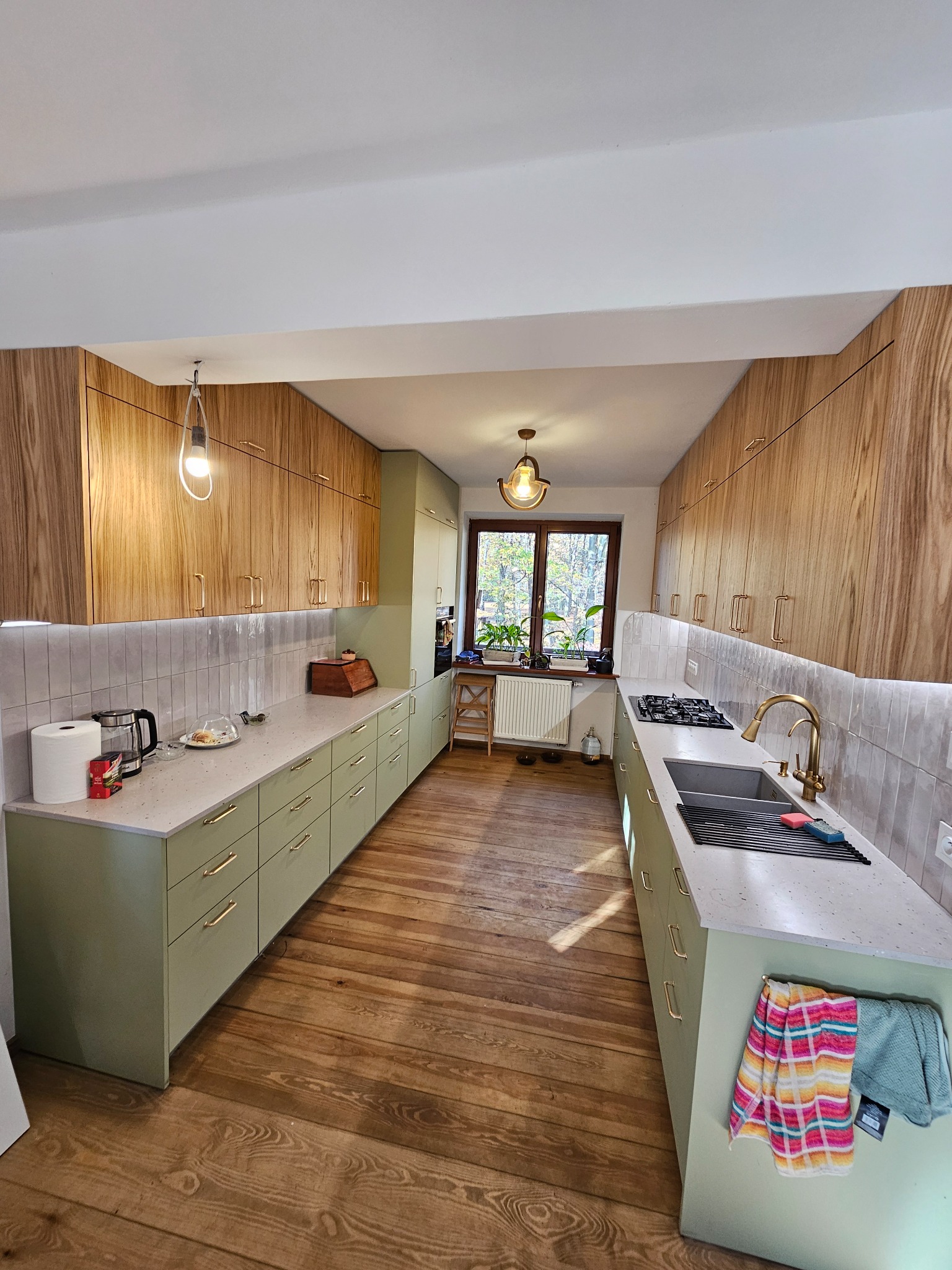 Wąska kuchnia z drewnianymi szafkami górnymi i zielonymi dolnymi, blatem z jasnego kamienia, drewnianą podłogą i oknem z widokiem na drzewa.