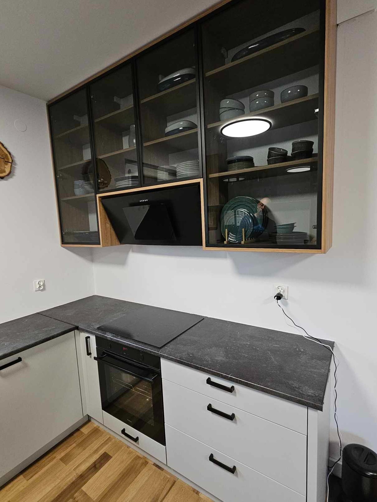 Nowoczesna kuchnia z białymi szafkami i blatem w odcieniach szarości, szklane szafki wiszące z oświetleniem LED eksponują naczynia, a czarny okap dodaje elegancji.