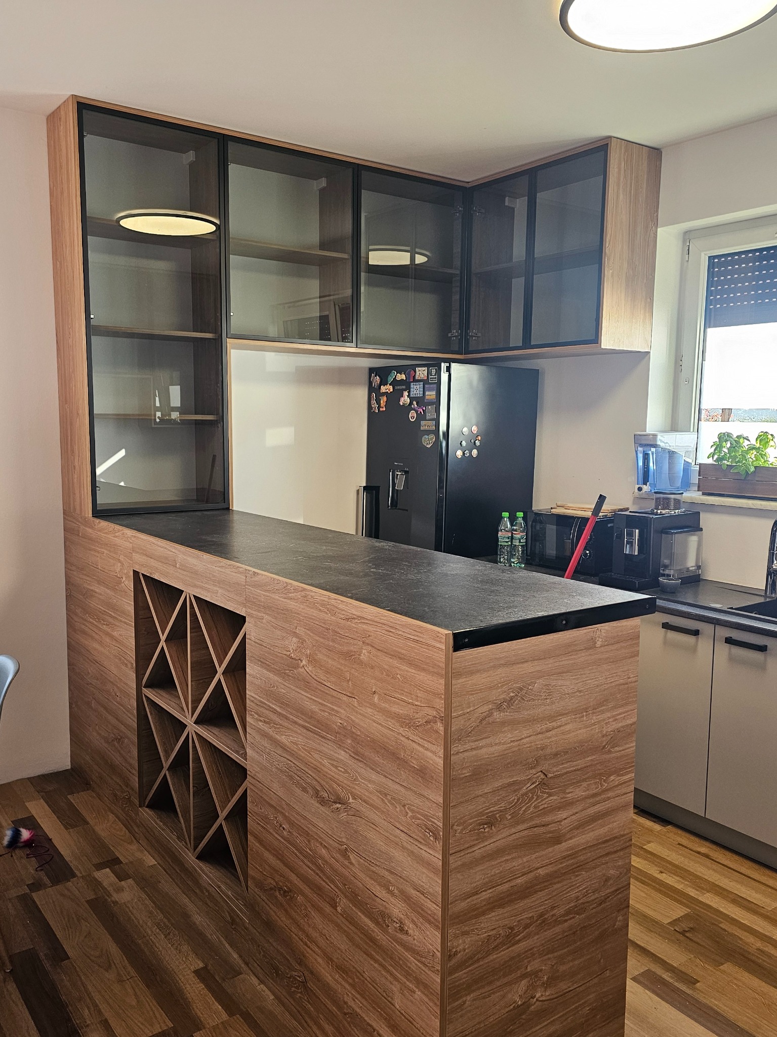 Nowoczesna kuchnia z drewnianą wyspą z wbudowanym regałem na wino, przeszklonymi szafkami wiszącymi z czarnymi ramami, czarnym blatem i sprzętem AGD w tle.