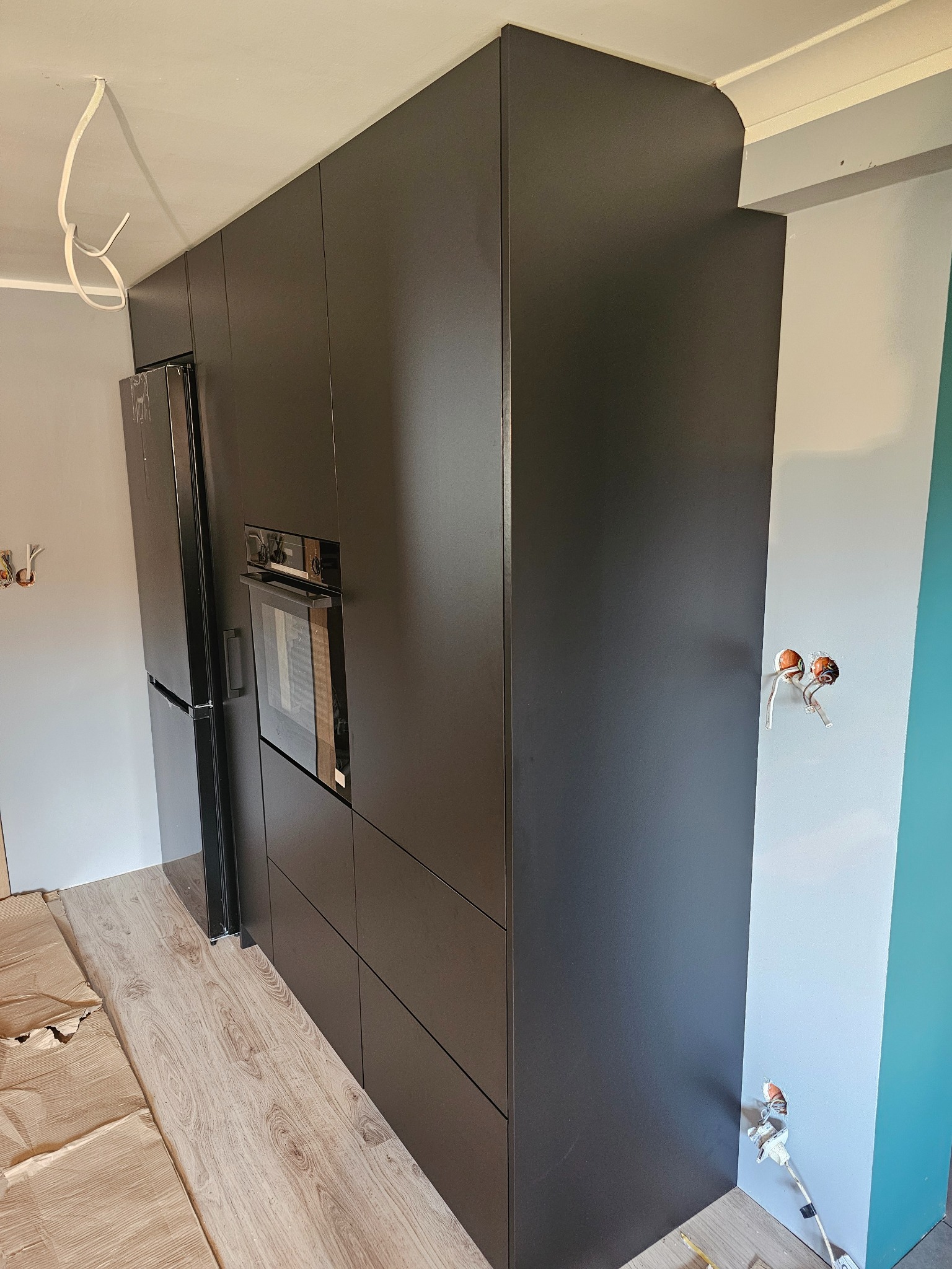 Ciemna zabudowa kuchenna z piekarnikiem i lodówką w trakcie montażu, widoczne wystające kable elektryczne i zabezpieczona podłoga.