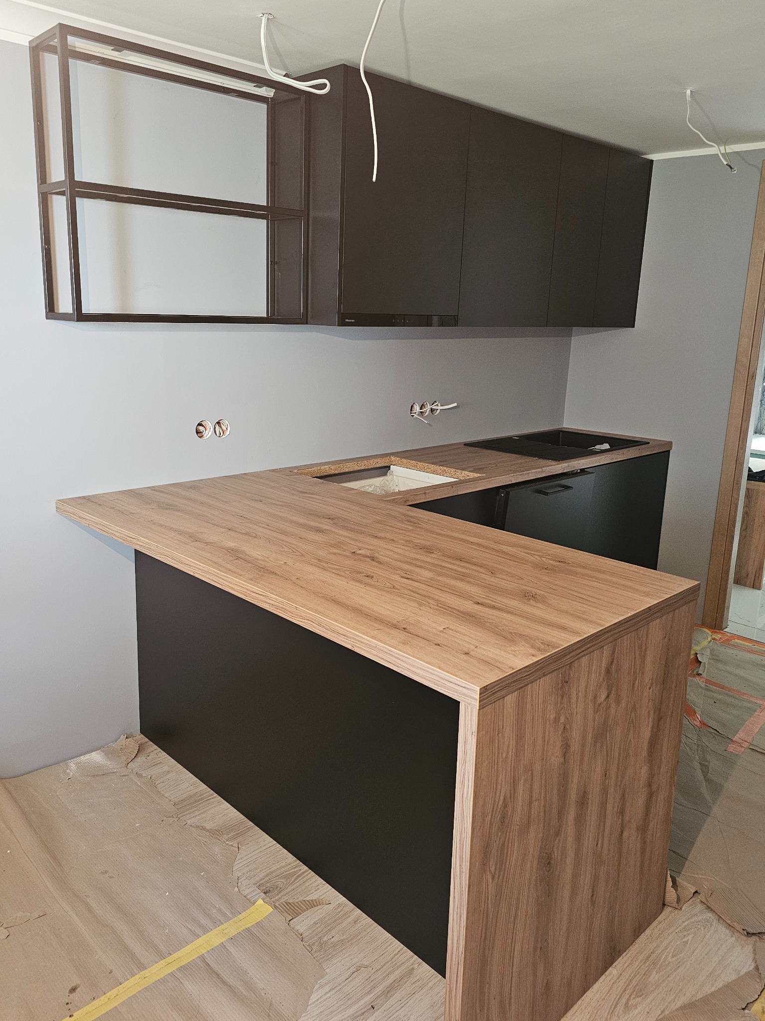 Częściowo zamontowana kuchnia z drewnianym blatem i czarnymi szafkami, widoczne niedokończone podłogi i wystające kable.