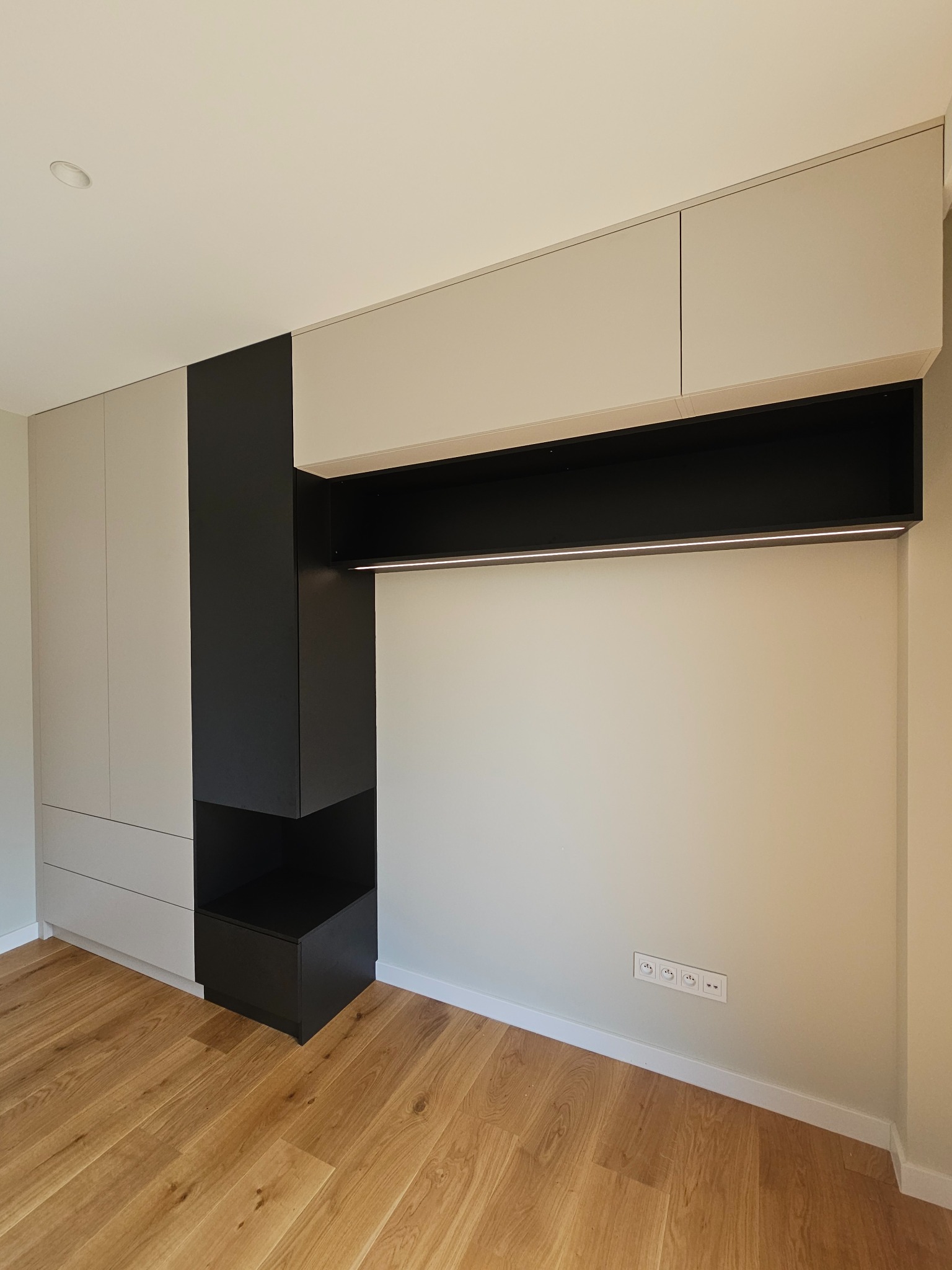 Nowoczesna zabudowa meblowa z szarymi szafkami i czarnym pionowym elementem, oświetlenie LED pod szafkami wiszącymi, parkiet na podłodze i gniazdka elektryczne na ścianie.