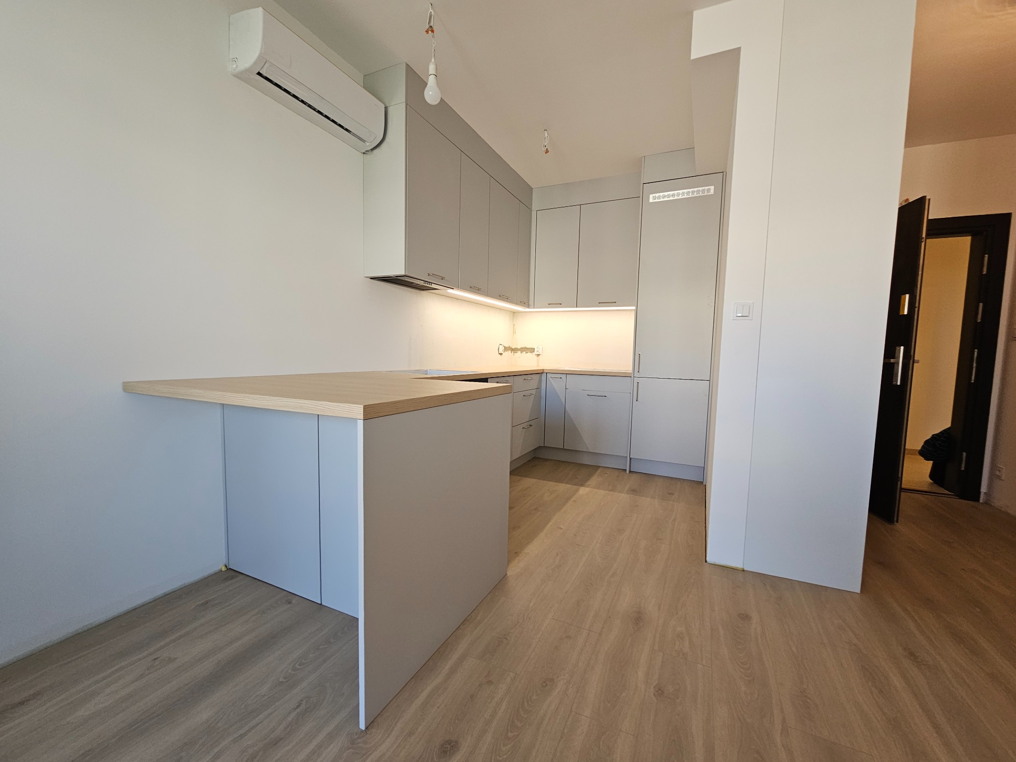 Jasna kuchnia z wyspą, szare fronty, drewniany blat. Widoczny klimatyzator na ścianie. Podłoga drewniana. Minimalistyczny design z otwartym przejściem do innego pomieszczenia.