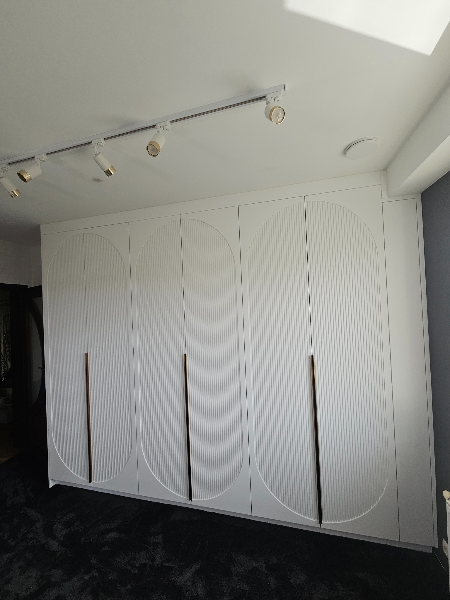 Biała szafa wnękowa z frezowanymi drzwiami w kształcie łuków, z podłużnymi uchwytami, oświetlona reflektorami na szynie. Nowoczesny design.