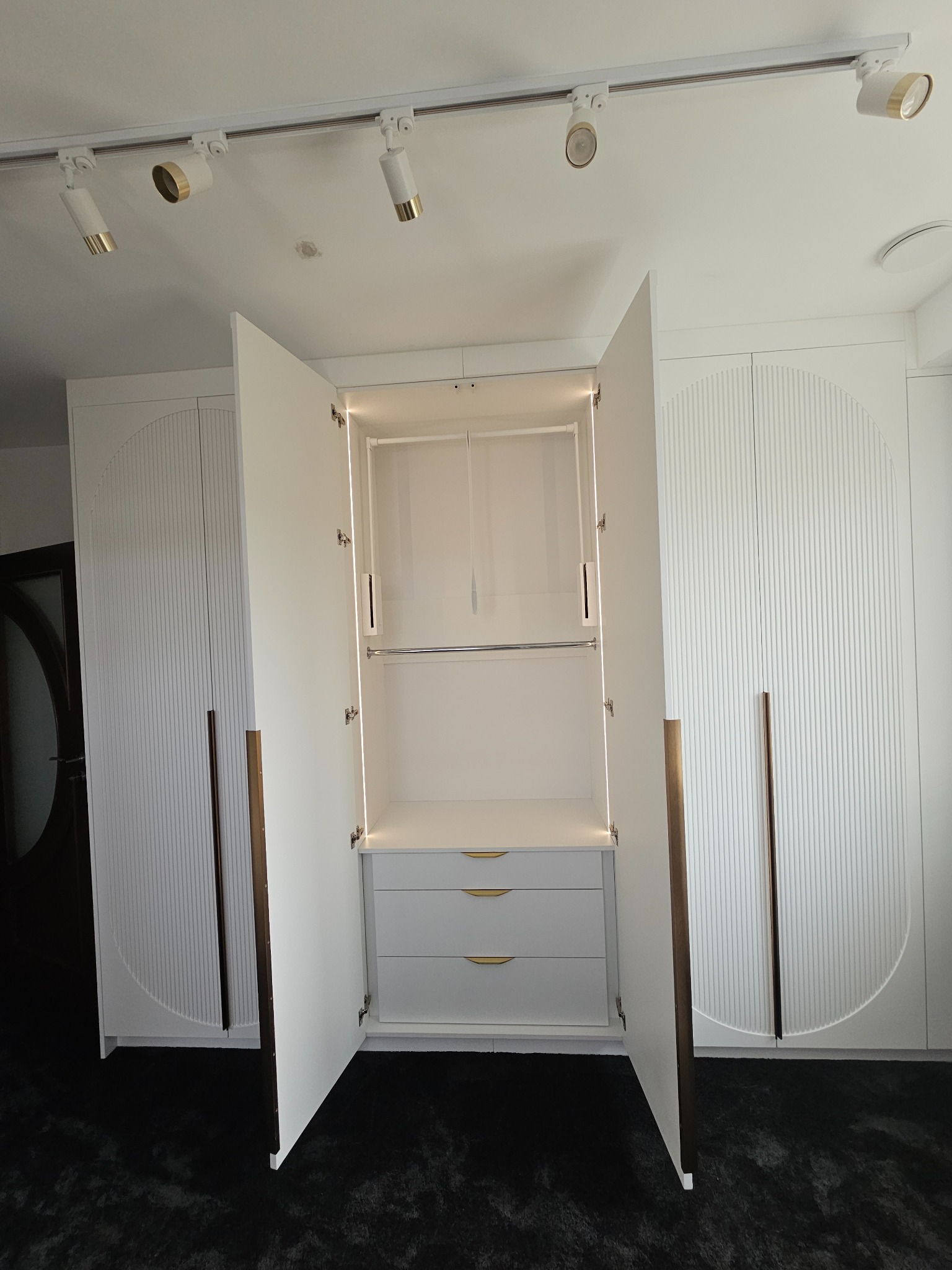 Nowoczesna biała szafa wnękowa z dekoracyjnymi frezami i złotymi uchwytami, oświetlenie LED wewnątrz, otwarte drzwi.