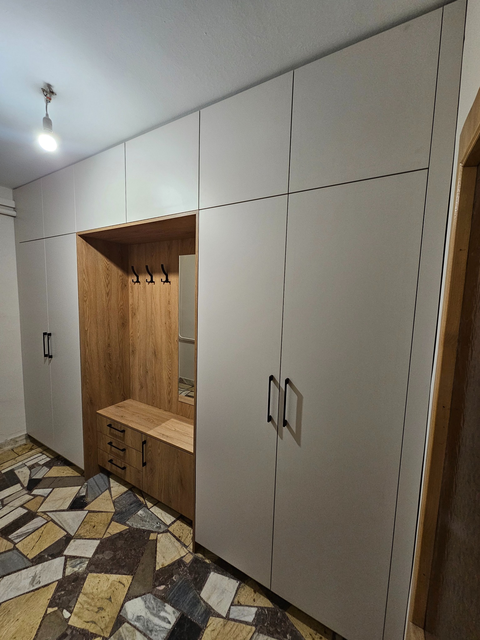 Jasnoszara szafa wnękowa z drewnianym wykończeniem i lustrem w przedpokoju w Pszczynie. Widoczne czarne, podłużne uchwyty i wzorzysta podłoga.