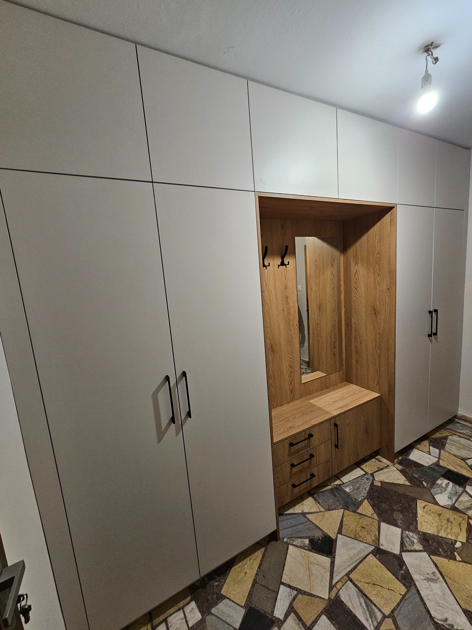 Nowoczesna szafa wnękowa z drewnianym wykończeniem i wbudowanym siedziskiem, uzupełniona o lustro i wieszaki, na tle wzorzystej podłogi.