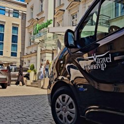 Poland VIP Travel - Czarny bus z napisem 'Vtravel Poland Vip' zaparkowany na brukowanym chodniku przed hotelem Holiday Inn, w tle widoczni piesi.