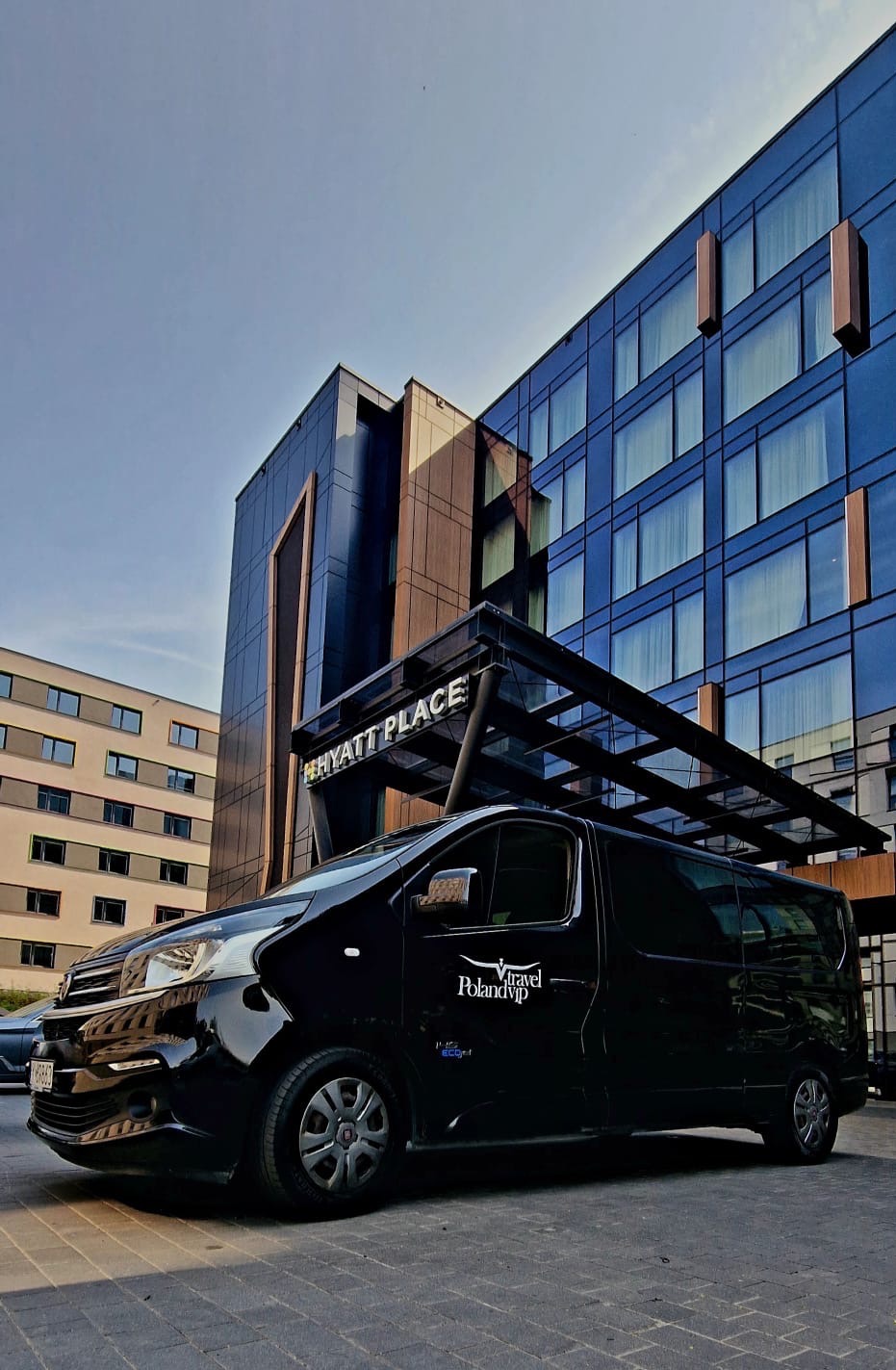 Czarny bus z logo PolandVIP Travel zaparkowany przed hotelem Hyatt Place, błękitne niebo w tle.