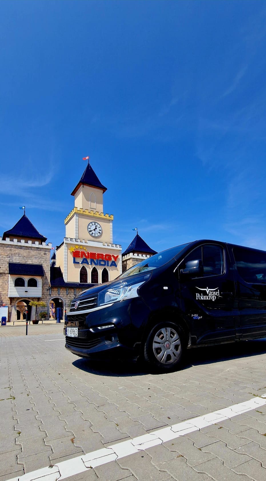 Czarny bus z logo Polandvip Travel na tle parku rozrywki Energylandia w Zatorze, zaparkowany na kostce brukowej w słoneczny dzień.