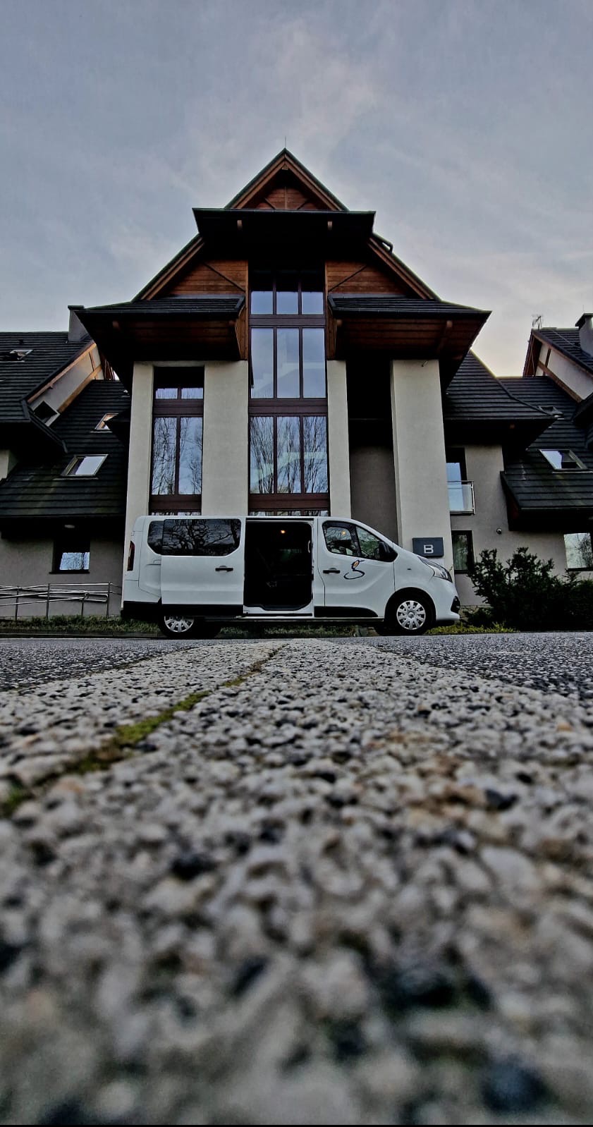 Biały bus z otwartymi drzwiami stoi przed budynkiem w stylu góralskim, widok z niskiej perspektywy na kamienną drogę.