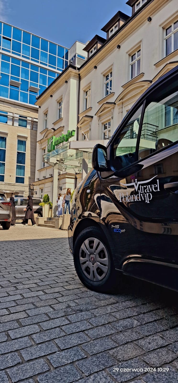 Czarny bus z napisem 'Vtravel Poland Vip' zaparkowany na brukowanym chodniku przed hotelem Holiday Inn, w tle widoczni piesi.
