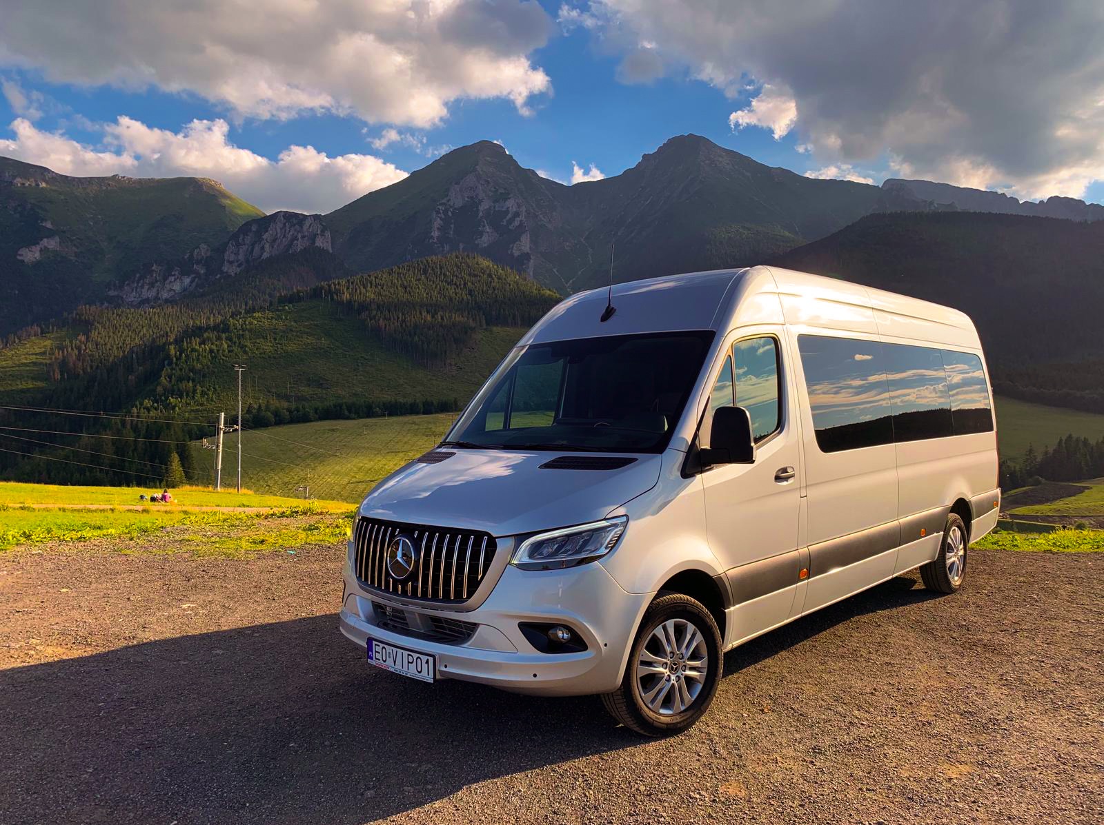 Srebrny bus Mercedes-Benz Sprinter z widokiem na Tatry w tle, oświetlony popołudniowym słońcem, na pierwszym planie żwirowa droga.