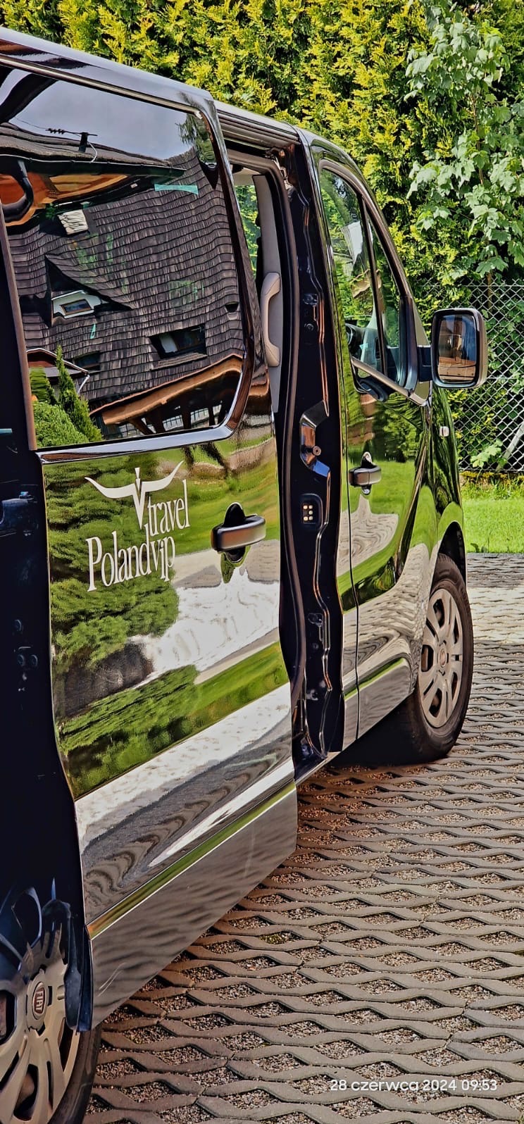 Czarny bus z otwartymi drzwiami, na karoserii widoczne logo 'Travel Polandvip', odbijające się otoczenie: budynek i zieleń.