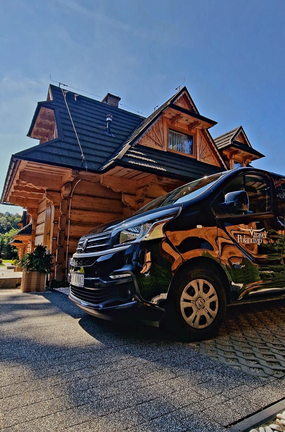 Czarny bus z logiem Poland VIP Travel zaparkowany przed drewnianym domem w stylu góralskim, widoczny numer rejestracyjny pojazdu.