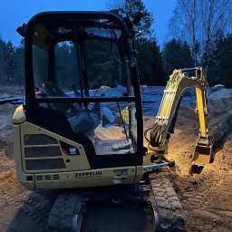 Budwest - Żółta minikoparka CAT 301.7D Zeppelin w trakcie pracy, zagłębiona łyżką w wykopie ziemnym, w tle widoczne drzewa i niebo o zmierzchu.