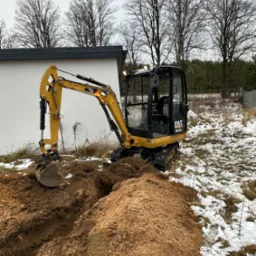 Żółta minikoparka CAT 301.7D kopie wąski rów w ziemi, obok leży pryzma wykopanej ziemi, na ziemi resztki śniegu, w tle budynek i drzewa.