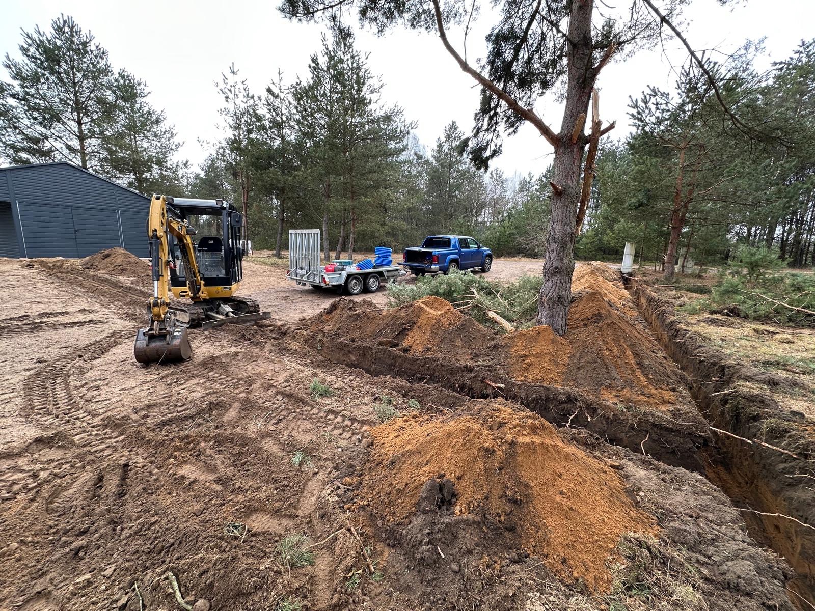 Żółta minikoparka CAT przy wykopanym rowie na piaszczystym terenie, w tle przyczepa z niebieskimi zbiornikami i niebieski pickup, obok las sosnowy i budynek gospodarczy.