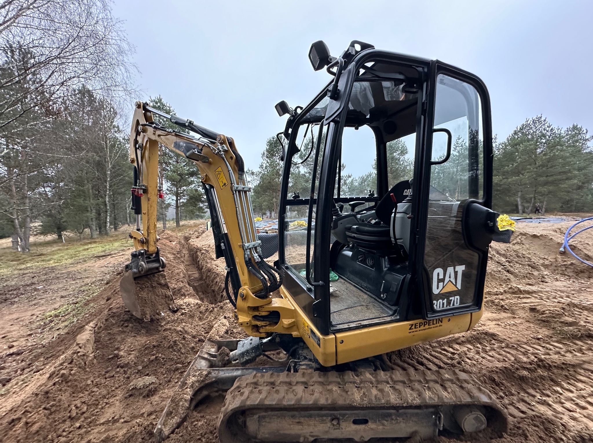 Żółta minikoparka CAT 301.7D z otwartą kabiną, widoczny wykopany wąski rów w piaszczystej ziemi, w tle drzewa i pochmurne niebo.
