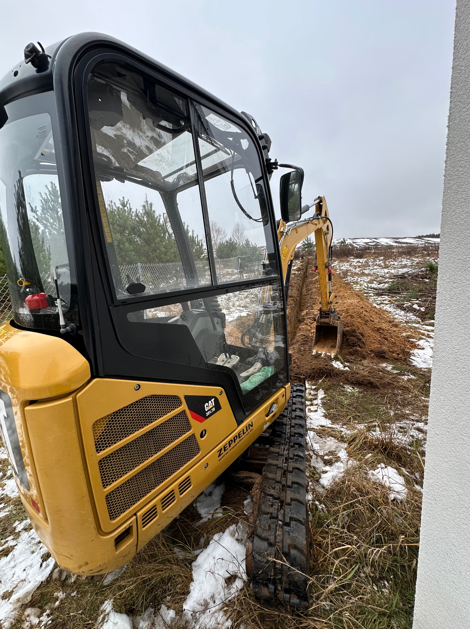 Żółta minikoparka CAT 301.7D w trakcie kopania wąskiego rowu na zaśnieżonym terenie, widok z boku kabiny.
