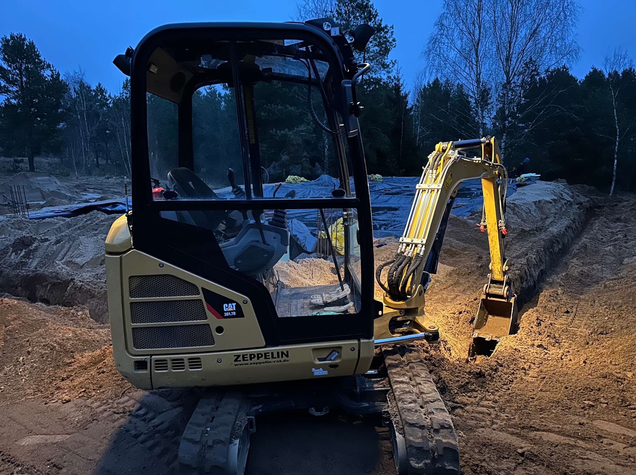 Żółta minikoparka CAT 301.7D Zeppelin w trakcie pracy, zagłębiona łyżką w wykopie ziemnym, w tle widoczne drzewa i niebo o zmierzchu.