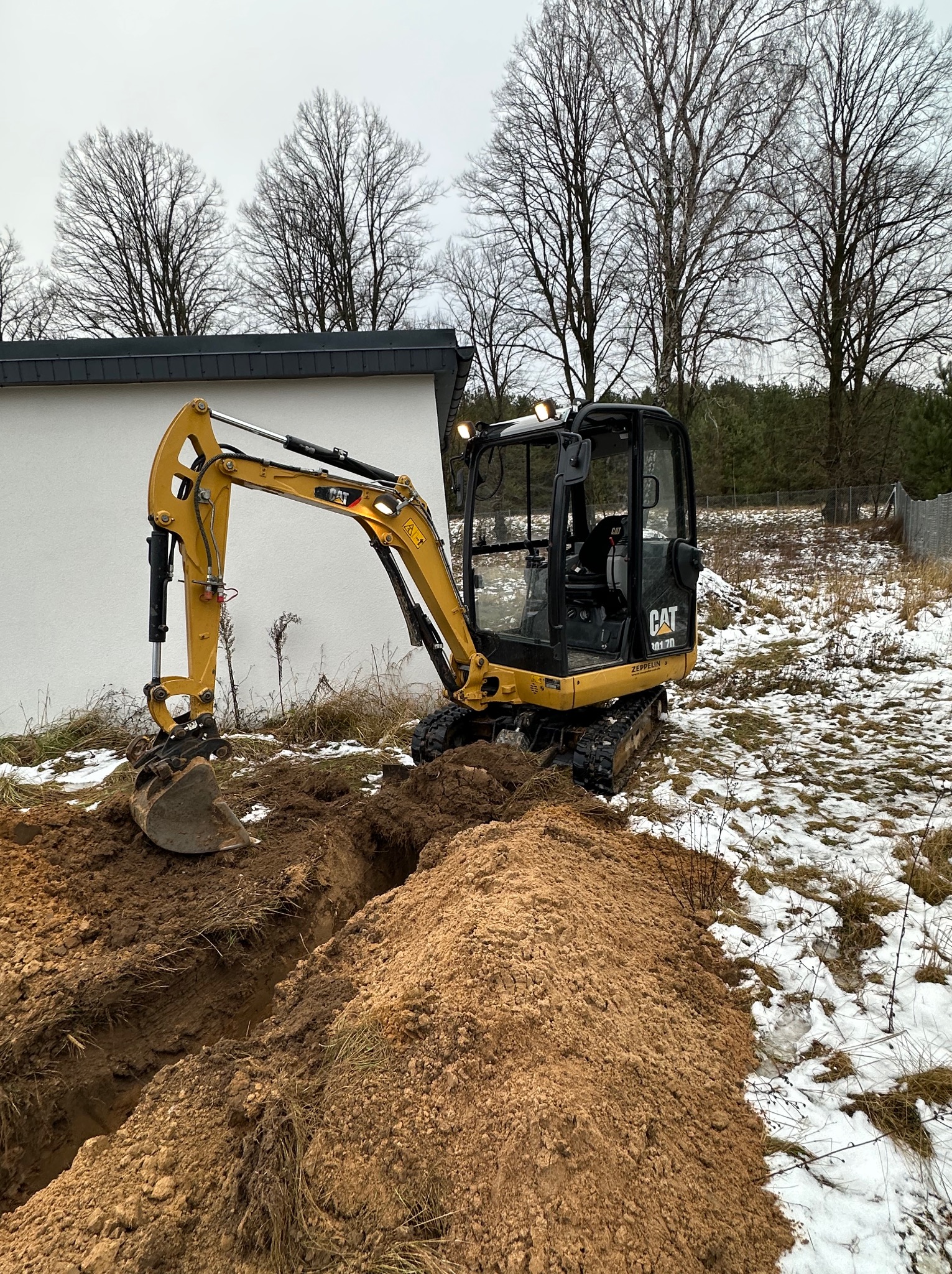 Żółta minikoparka CAT 301.7D kopie wąski rów w ziemi, obok leży pryzma wykopanej ziemi, na ziemi resztki śniegu, w tle budynek i drzewa.