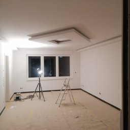 PPHU Jedynka - Pomalowane na biało wnętrze pokoju z gładzią na ścianach i ozdobnym sufitem podwieszanym z oświetleniem, zabezpieczona podłoga, drabina i lampa budowlana na trójnogu.