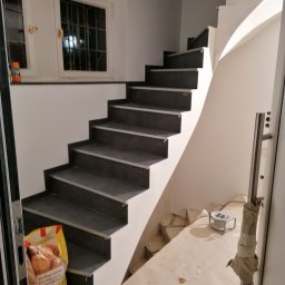 PPHU Jedynka - Klatka schodowa z ciemnymi stopnicami i białymi podstopnicami w trakcie remontu, widoczny kabel przedłużający na podłodze i torba z zakupami na schodach.