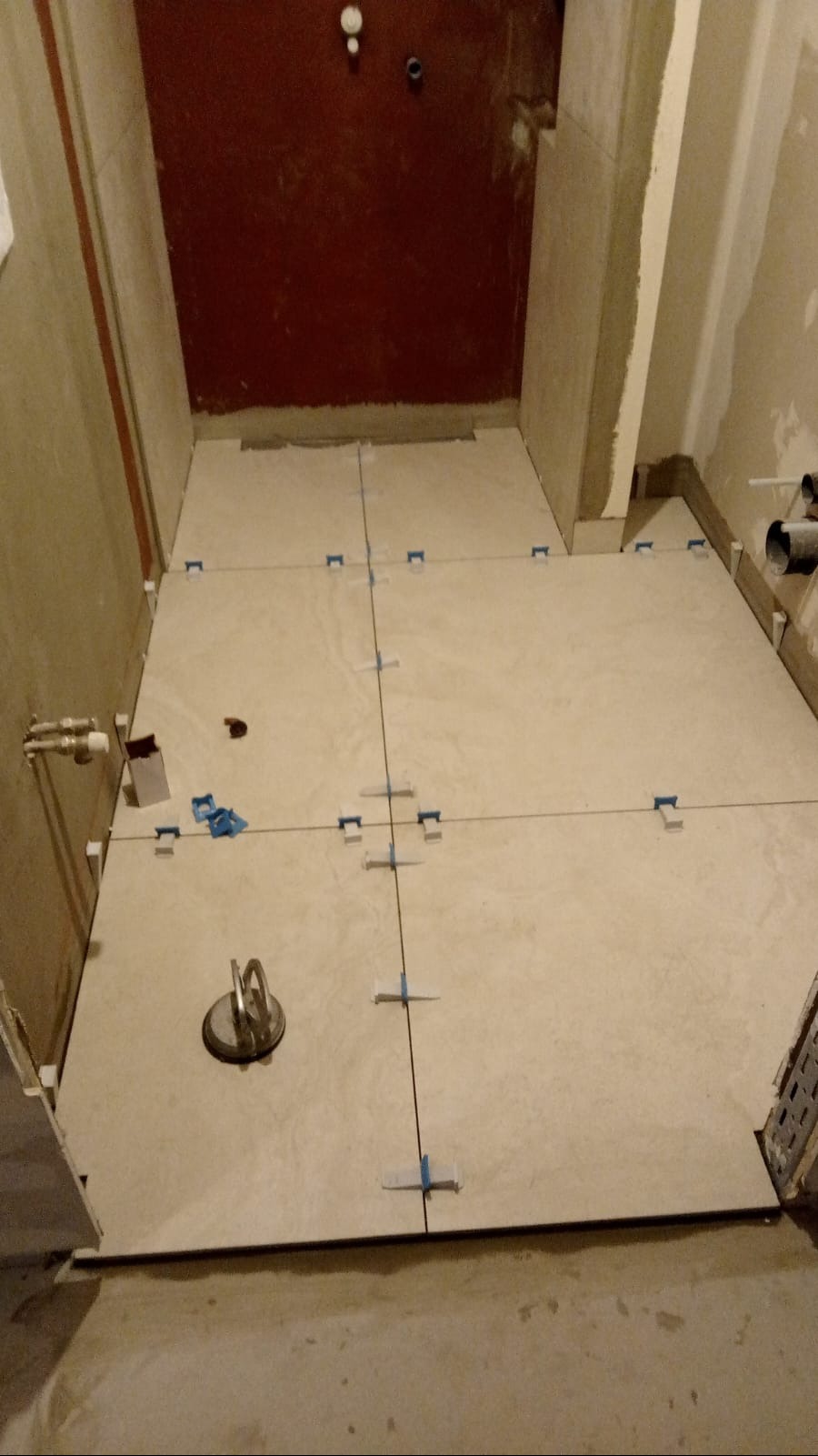 Podłoga w łazience w trakcie układania płytek, widoczne krzyżyki dystansowe, przyssawka do płytek i zaznaczone linie cięcia. Czerwona ściana w tle.