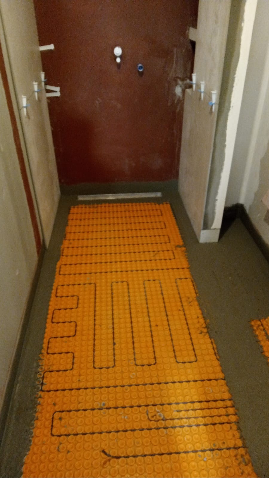 Pomarańczowa mata grzewcza ułożona na podłodze w trakcie prac wykończeniowych łazienki, widoczne ściany przygotowane do położenia płytek.