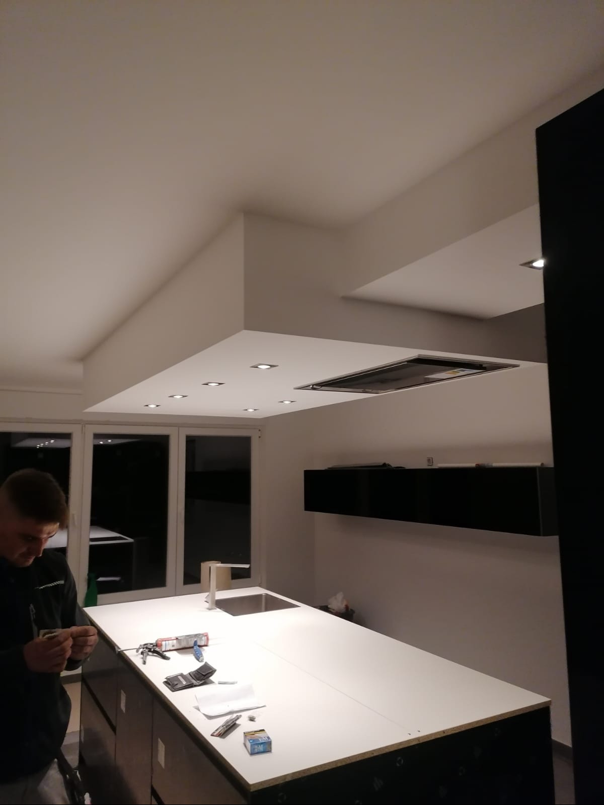 Montaż oświetlenia podszafkowego w nowoczesnej kuchni z wyspą, widoczny monter przy pracy i narzędzia na blacie.