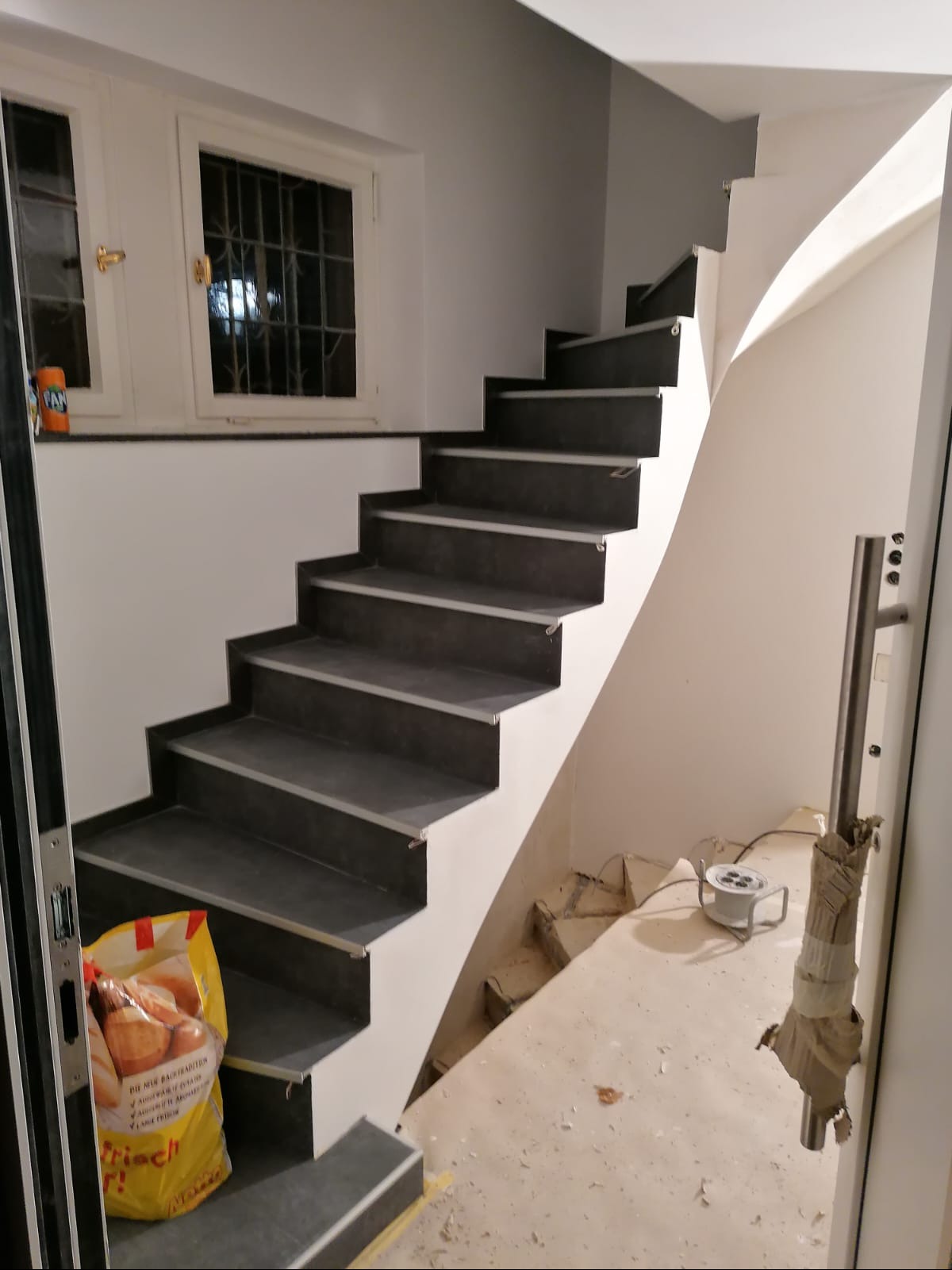 Klatka schodowa z ciemnymi stopnicami i białymi podstopnicami w trakcie remontu, widoczny kabel przedłużający na podłodze i torba z zakupami na schodach.