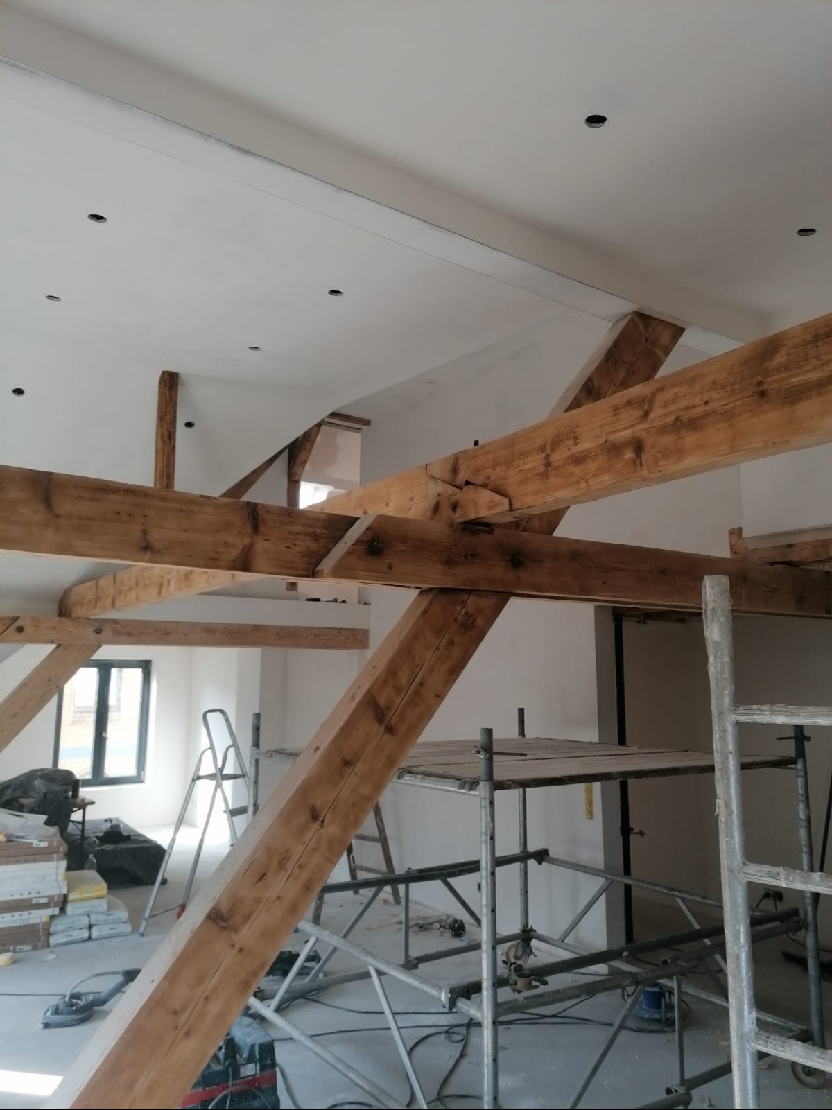Wnętrze w trakcie renowacji z widocznymi drewnianymi belkami konstrukcyjnymi, rusztowaniami, drabiną i workami z materiałami budowlanymi w tle, oświetlenie punktowe w suficie.