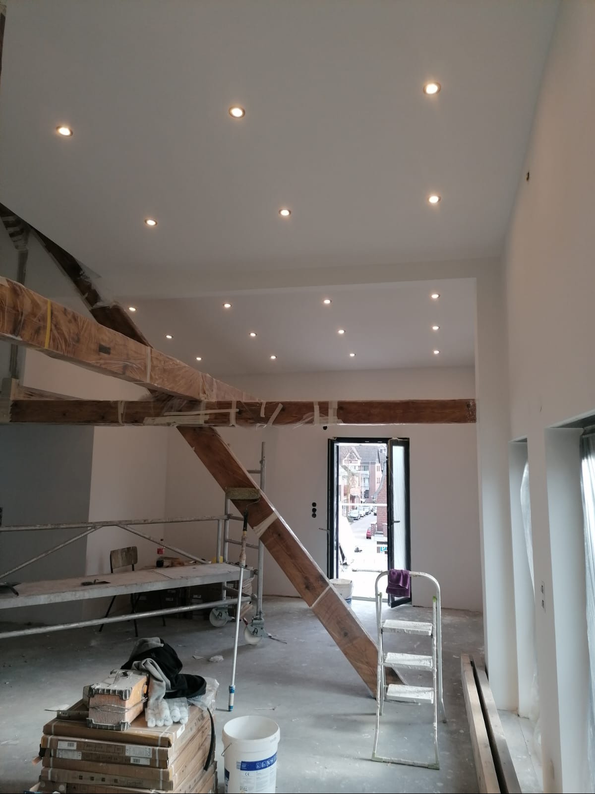 Wnętrze w trakcie remontu z odsłoniętymi drewnianymi belkami zabezpieczonymi folią, świeżo pomalowanymi ścianami i sufitem z wbudowanymi punktowymi lampami, widoczne rusztowania i drabina, w tle...