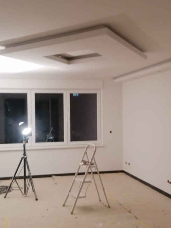 Wnętrze pokoju w trakcie prac wykończeniowych: białe ściany, sufit z dekoracyjną konstrukcją gipsową, drabina i lampa na statywie.