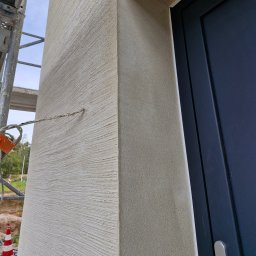 Wójcik tynki - Fragment elewacji budynku z nałożonym tynkiem strukturalnym w odcieniach beżu, widoczny narożnik i fragment ościeżnicy drzwi, rusztowanie w tle.