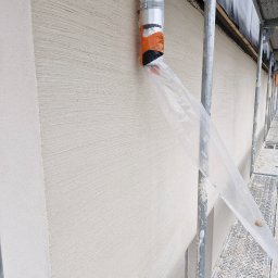 Wójcik tynki - Świeżo nałożona, jasna struktura na elewacji budynku widoczna ze stojącego rusztowania, z rurą odprowadzającą pył przymocowaną do ściany taśmą pomarańczową.