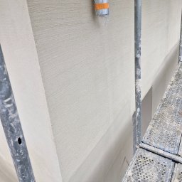 Wójcik tynki - Świeżo nałożony, jasny tynk zewnętrzny z widoczną fakturą, fragment rusztowania z przodu, rura wentylacyjna przy górnej krawędzi ściany.