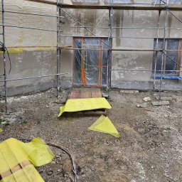 Wójcik tynki - Elewacja budynku w trakcie ocieplania, widoczne rusztowania, okna zabezpieczone folią, częściowo nałożony tynk, teren budowy z gruzem i żółtą siatką zabezpieczającą.