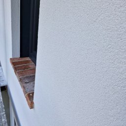 Wójcik tynki - Fragment elewacji budynku po nałożeniu białego, baranka, widoczne obramowanie okna z ciemnego tworzywa i ceglany parapet.