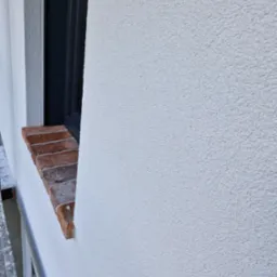 Fragment elewacji budynku po nałożeniu białego, baranka, widoczne obramowanie okna z ciemnego tworzywa i ceglany parapet.