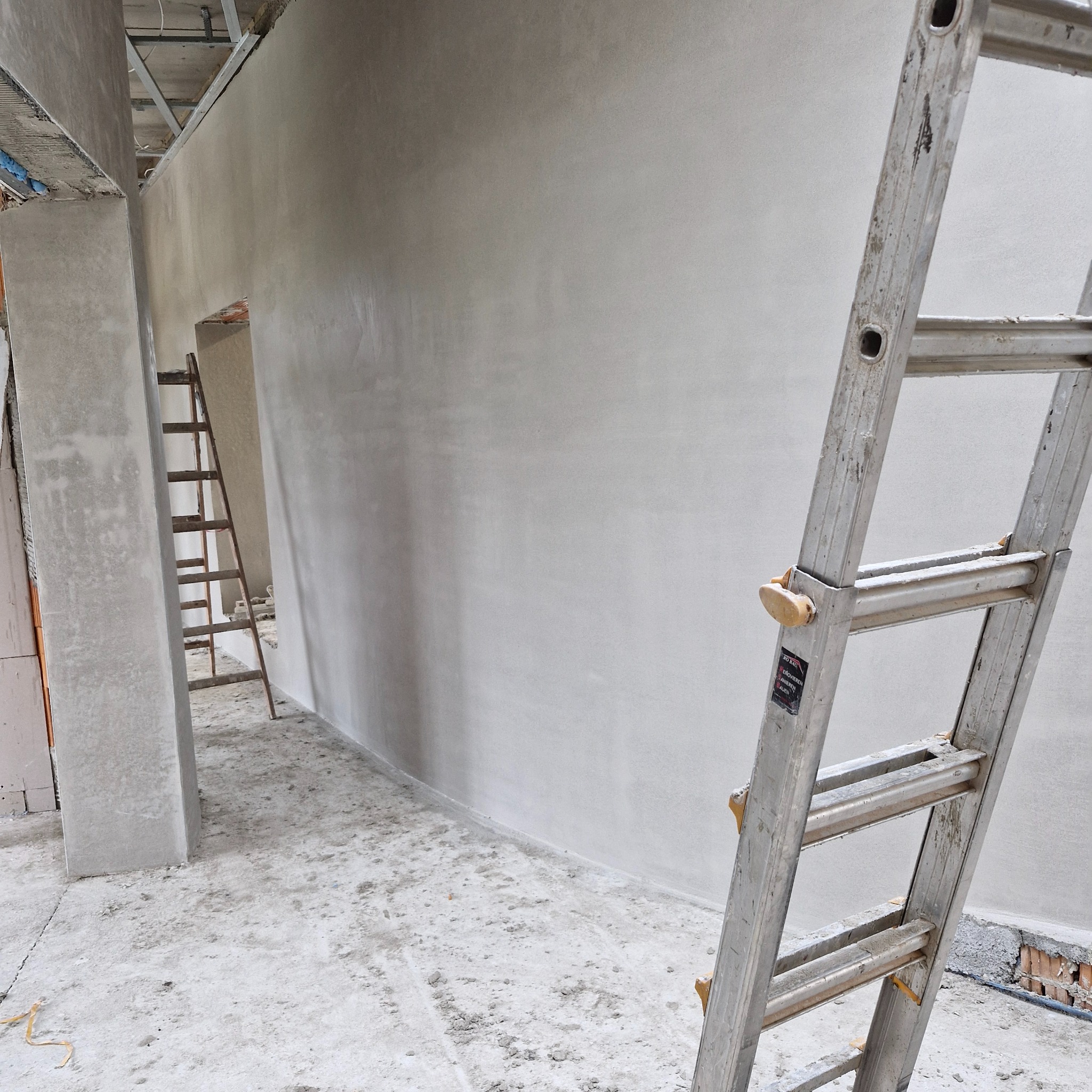 Świeżo nałożona, jasnoszara warstwa na ścianie w surowym wnętrzu, z widoczną drabiną aluminiową w pierwszym planie i drewnianą drabiną w tle, na podłodze resztki materiałów budowlanych.